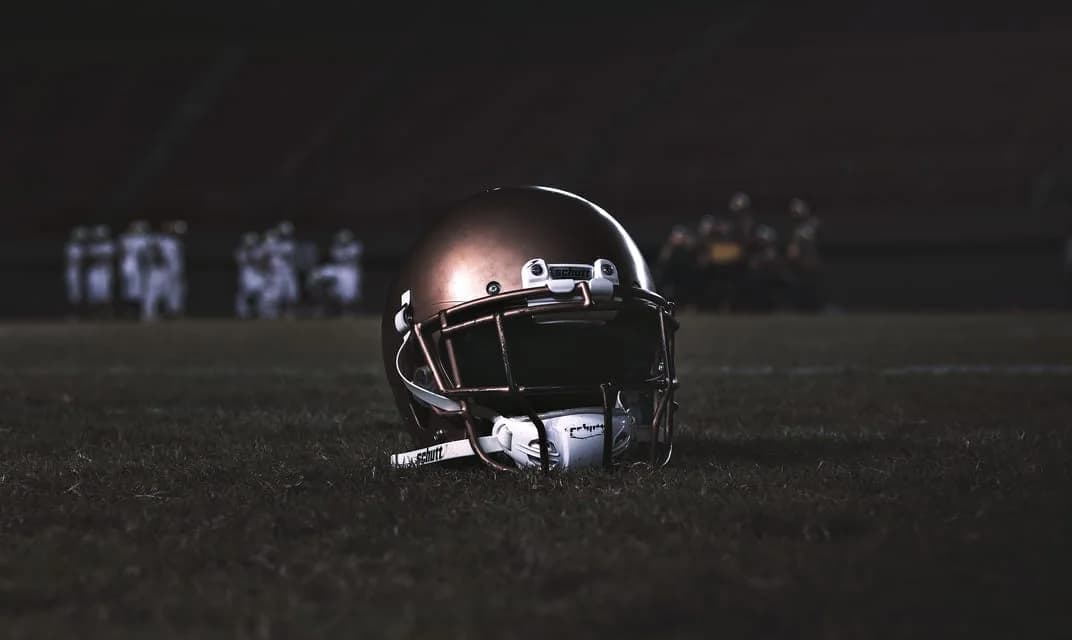 A football helmet resting on grass at dusk with players out of focus in the background.