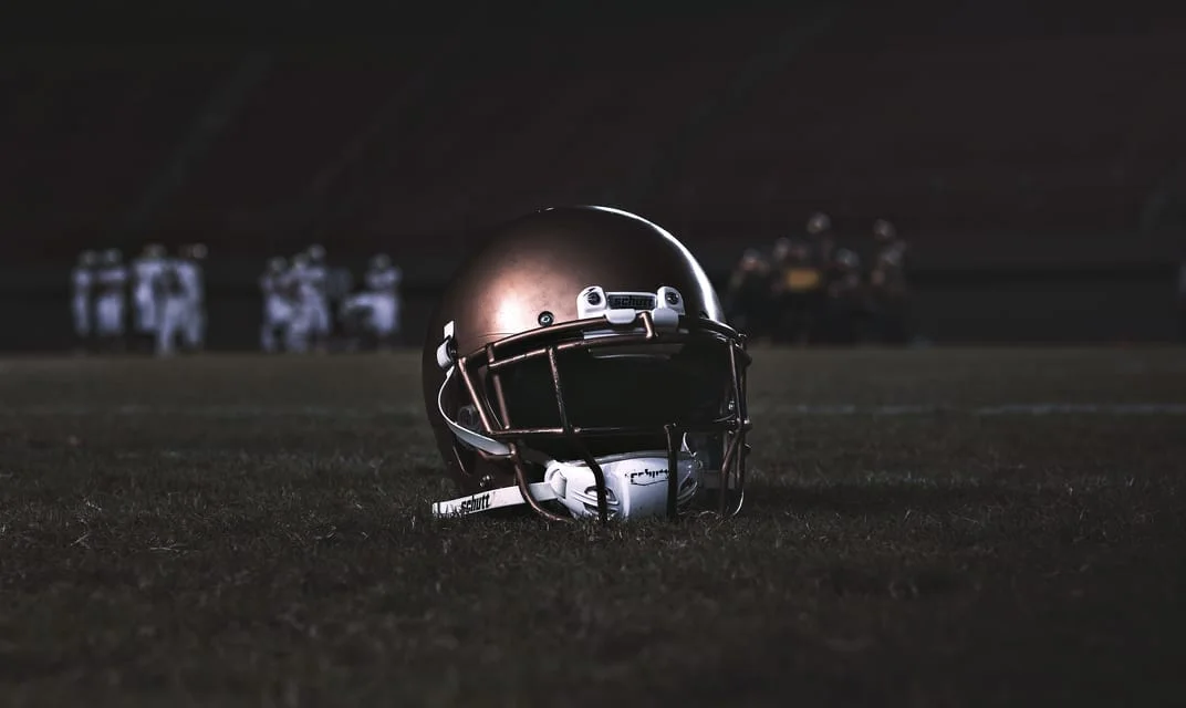 A football helmet resting on grass at dusk with players out of focus in the background.